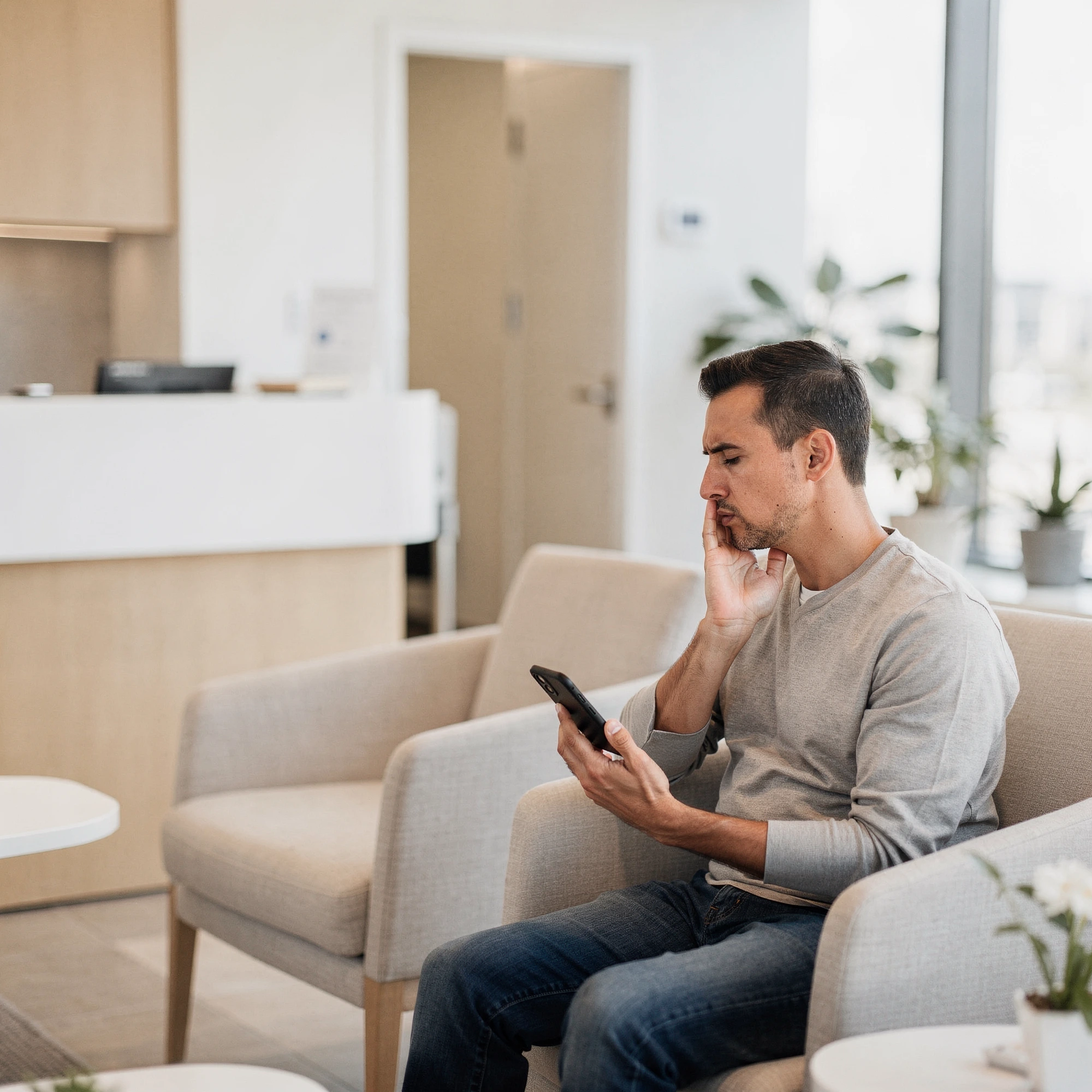 Man in waiting room holding phone with hand to face, appearing to have tooth pain, modern dental office in background